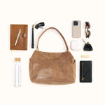 A brown leather shoulder bag is arranged in a styled flat lay with various accessories around it on a light cream background, including a notebook, pen, car key fob, phone case, sunglasses, earbuds case, wallet, and water bottle.