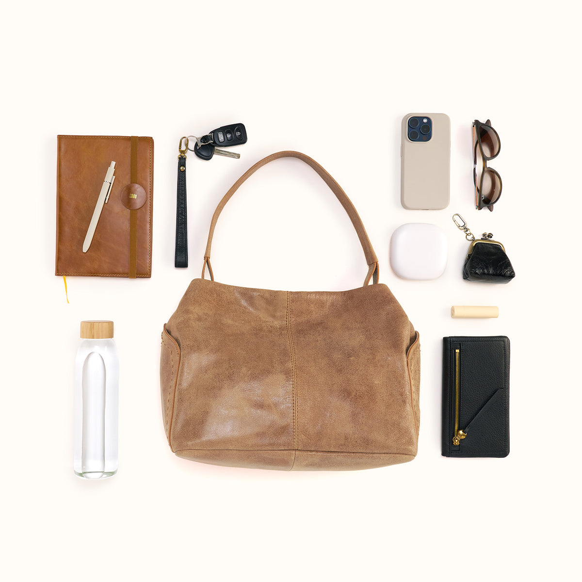 A brown leather shoulder bag is arranged in a styled flat lay with various accessories around it on a light cream background, including a notebook, pen, car key fob, phone case, sunglasses, earbuds case, wallet, and water bottle.