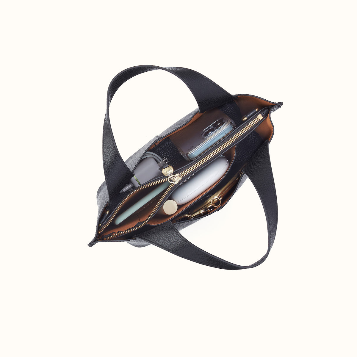 An overhead view of a black leather shoulder bag with its zipper open, showing its brown interior lining and compartments filled with accessories, on a solid off-white background.