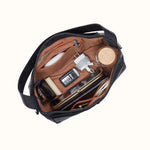 An open black shoulder bag with a tan interior, shown from above and filled with travel accessories including a white power adapter, a flip phone, a wooden lid, and a wallet, on a solid off-white background.
