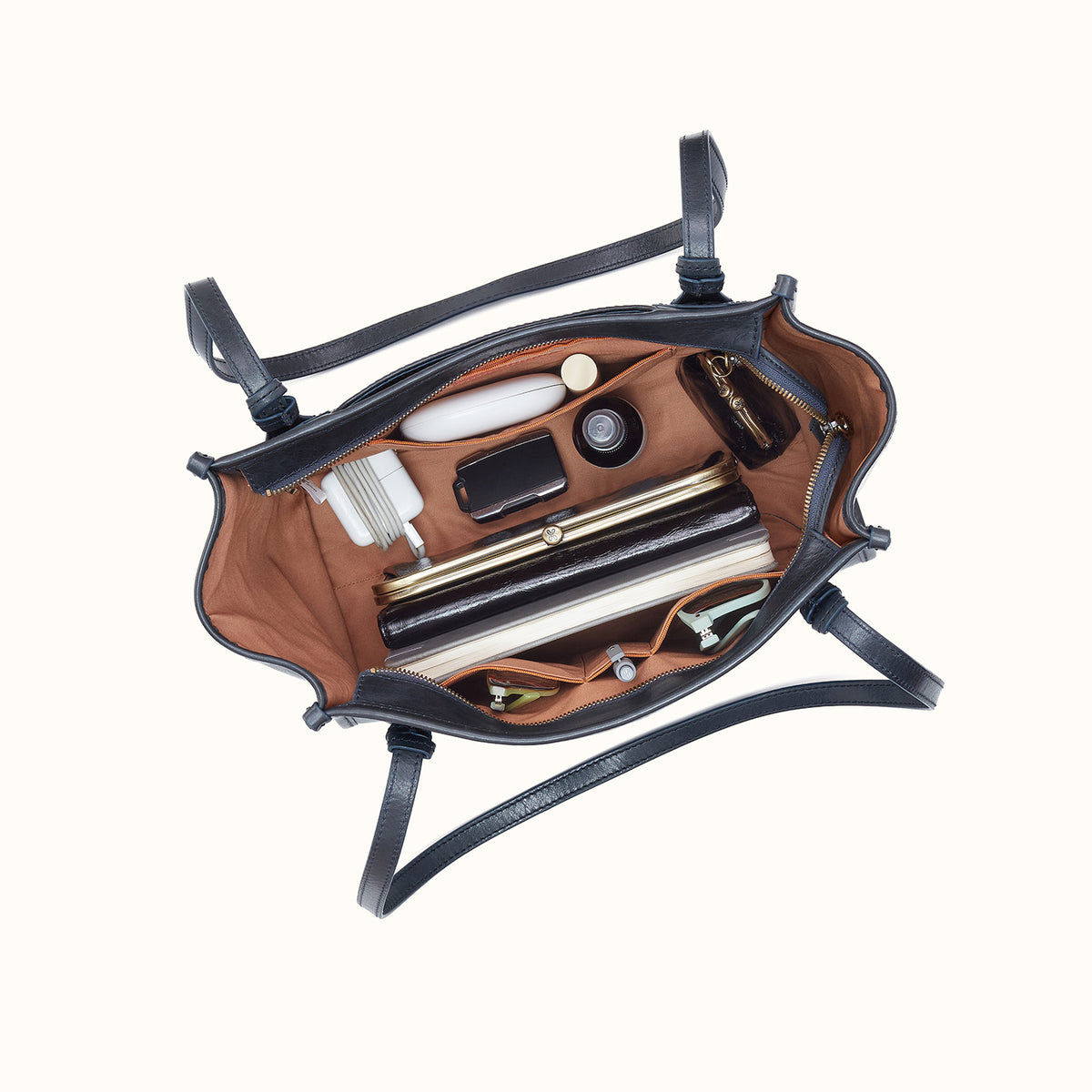 An overhead view of an open black leather handbag with a brown interior, shown packed with various cosmetics and accessories against a plain off-white background.