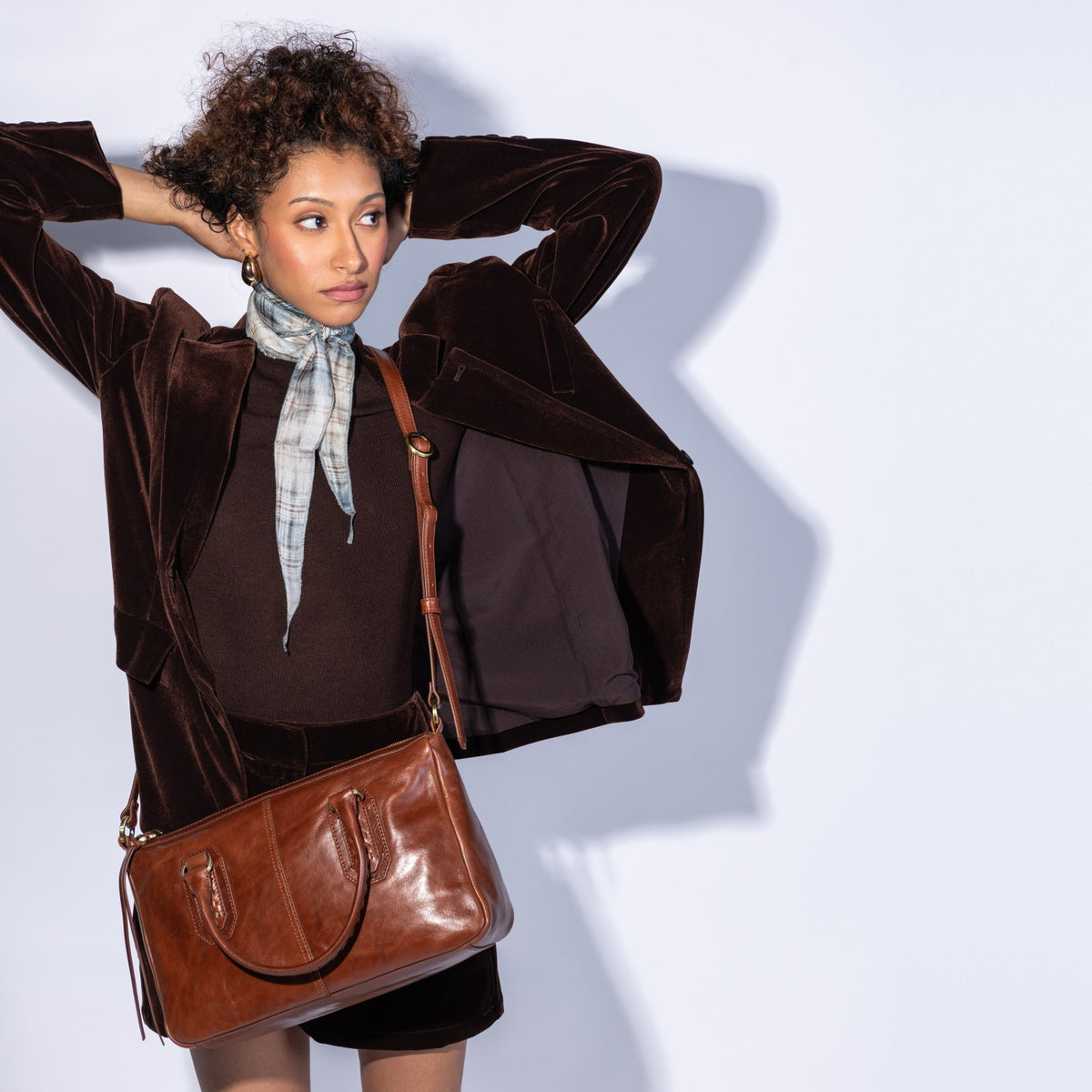 A woman poses in a dark brown velvet blazer, brown top, and a light blue patterned scarf, carrying a brown leather shoulder bag against a light gray background.