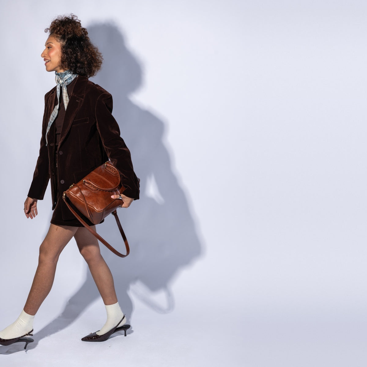 A woman walks in profile wearing a dark brown velvet blazer, a short skirt, white socks, and black heels while carrying a brown leather shoulder bag against a light gray studio background with a strong cast shadow.