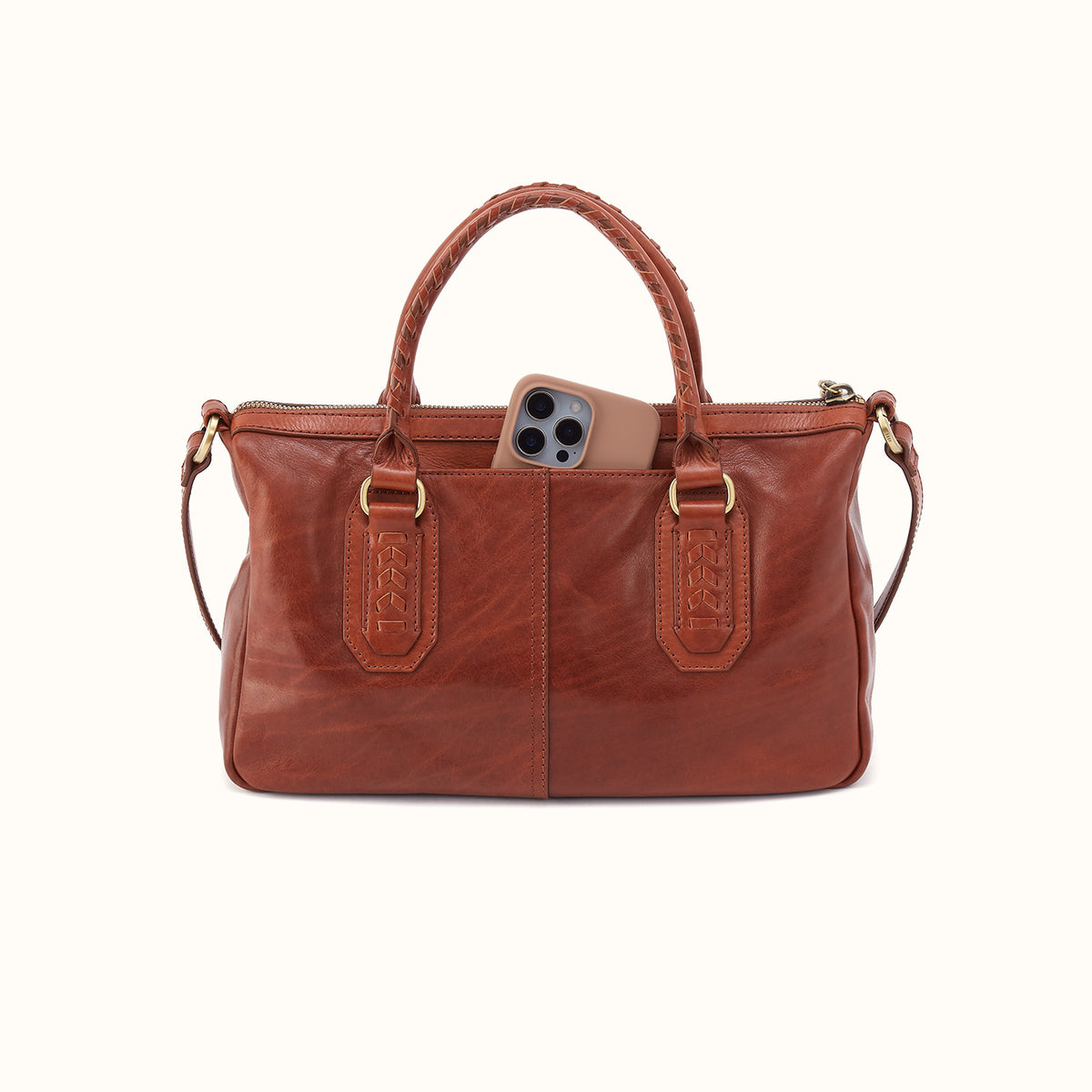 A brown leather handbag with braided top handles, gold-tone hardware, and a phone partially tucked into one of the front slip pockets, shown against a plain off-white background.