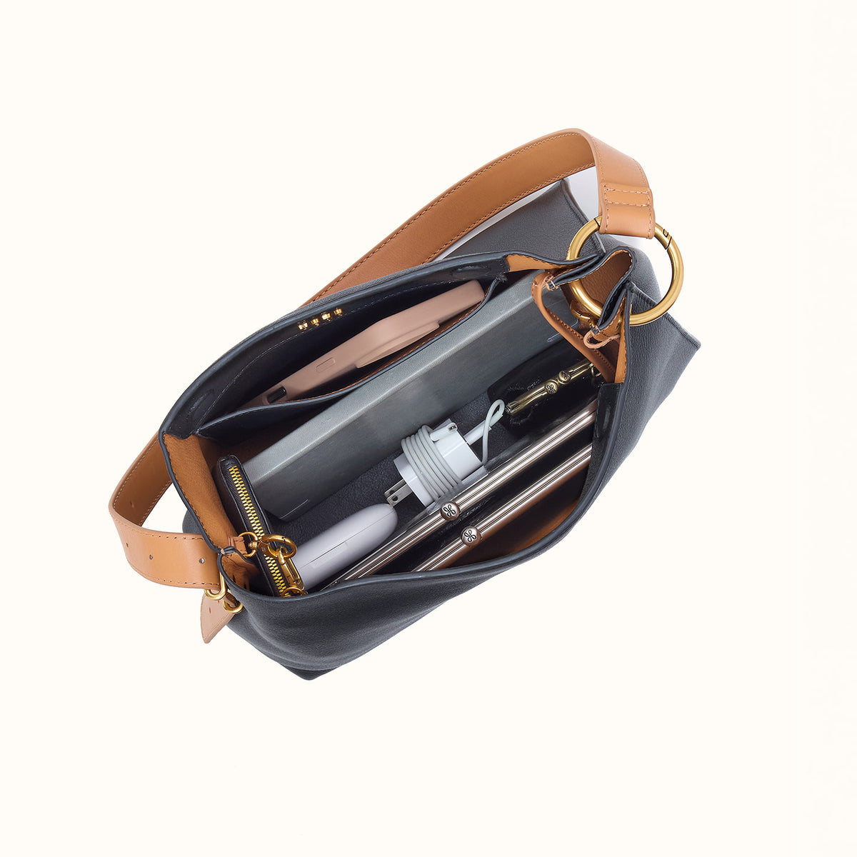 A top-down view of a black leather shoulder bag with tan leather trim and gold-tone hardware, shown open to reveal its interior compartments filled with travel accessories.