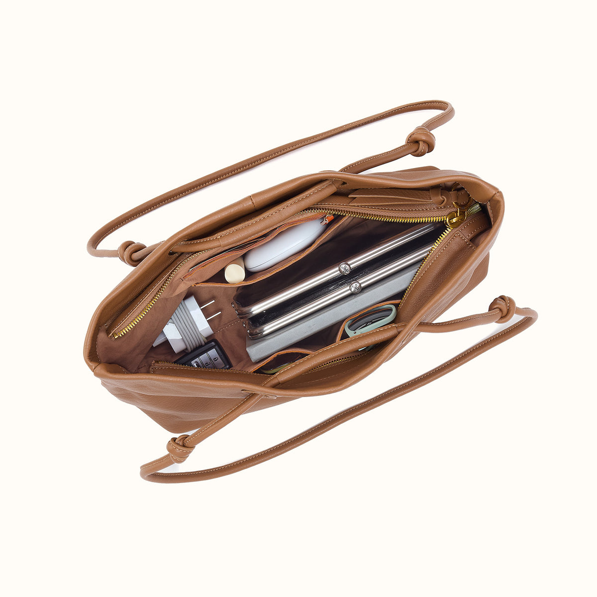 A tan leather shoulder bag shown open from a top-down view, revealing its interior compartments filled with makeup items such as pens, brushes, and a small bottle, on a plain off-white background.