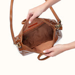 A tan leather handbag with a brown interior lining, shown open with a person's hands holding it against a plain off-white background.