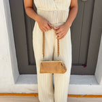 A person wearing a cream pleated jumpsuit holds a tan suede clutch with a woven brown strap in front of a gray wooden door.