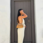 A woman in a cream pleated backless maxi dress stands in profile holding a tan leather shoulder bag with a braided strap, set against a dark gray wooden door and white walls.